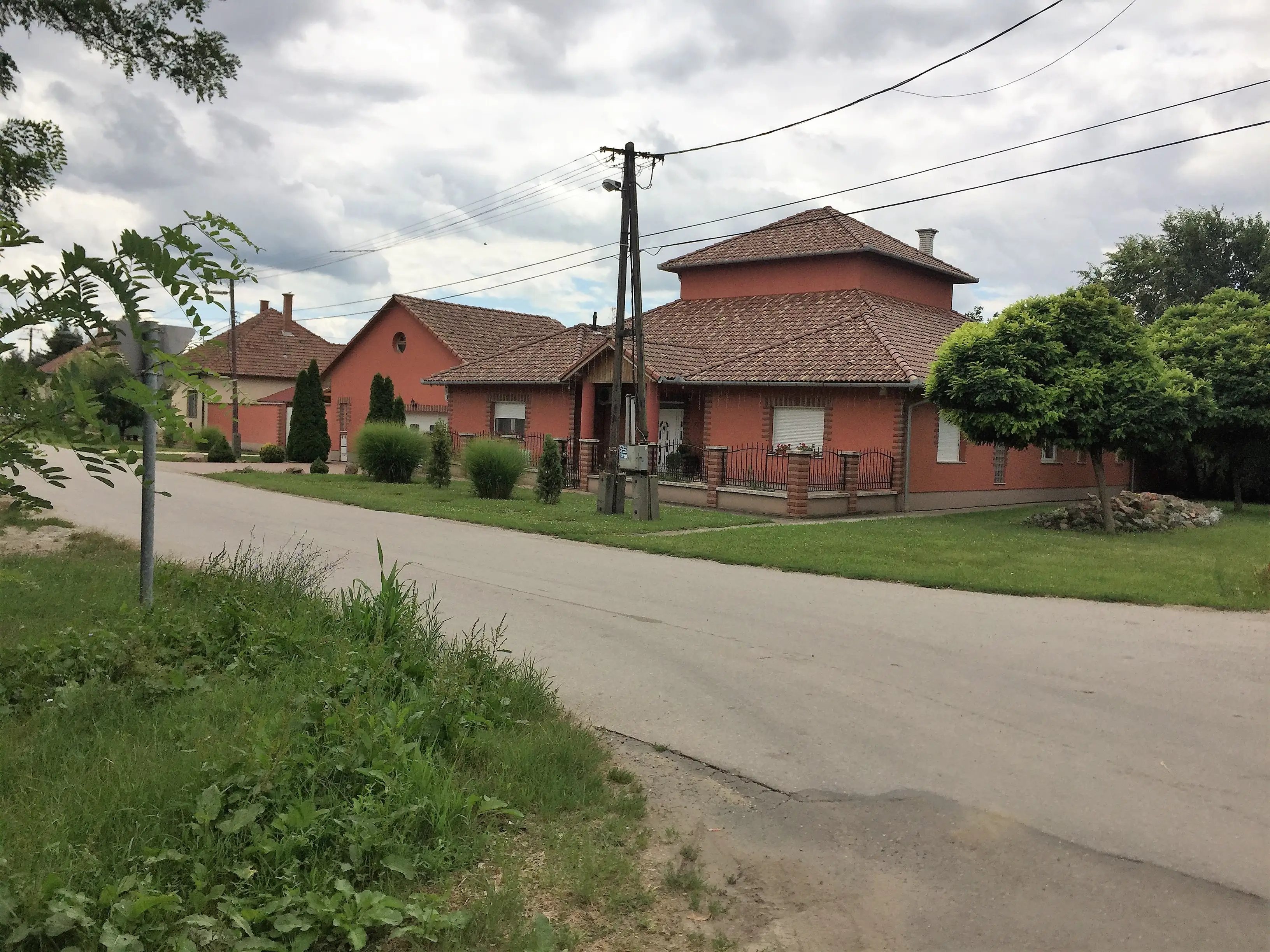 Vörös vakolatú, kétszintes sarokház sátortetővel egy kertes utcában